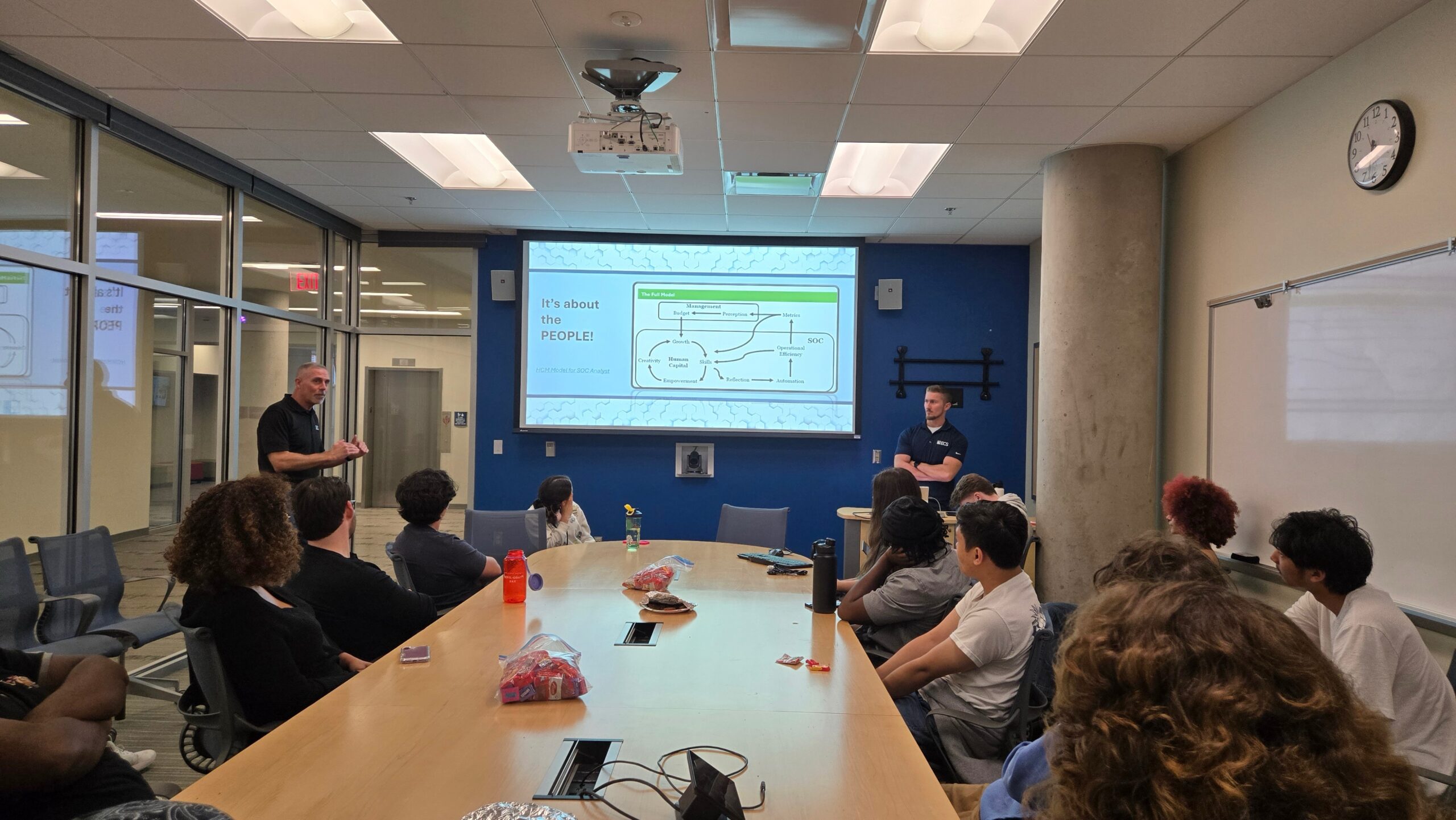 Members of UMW Cyber Club sitting around a conference table listening to two presenters speaking at the front of the room.